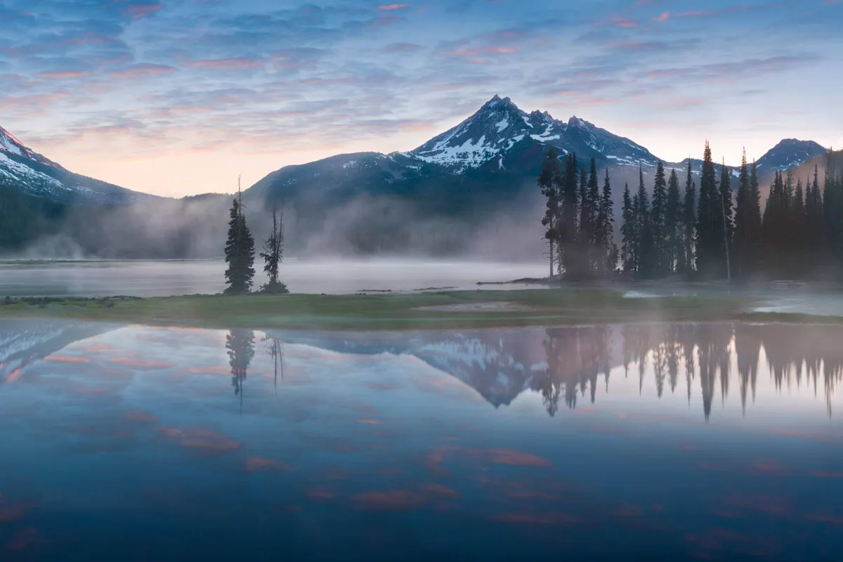 A tranquil view of misty mountains reflecting over a calm lake, surrounded by dense trees. The scene is bathed in soft natural light, evoking a sense of peace and connection with nature. The setting is perfect for outdoor activities like hiking and birdwatching, and offers a restorative escape.