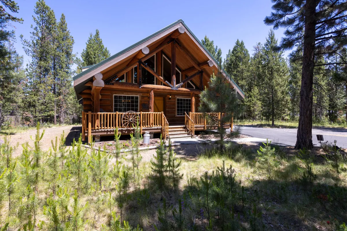 The image features a charming log cabin with rustic wood siding, nestled among tall pines in the tranquil Oregon high desert. A spacious wrap-around porch invites guests to relax, with a classic wooden wheel decoration adding to the cabin's cozy, welcoming vibe. The setting is serene, perfect for enjoying nature or gathering with loved ones.