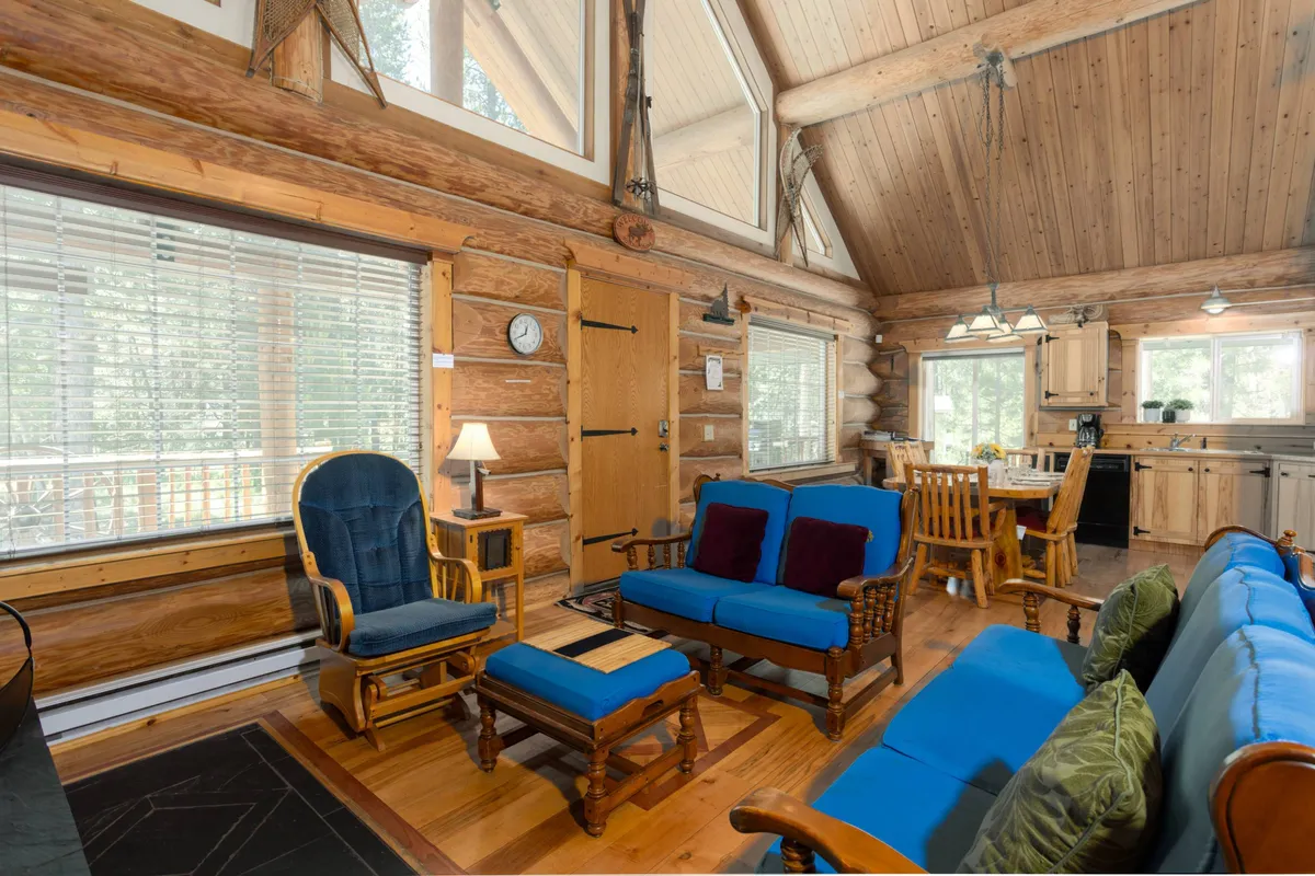 A welcoming indoor dining and living area with log walls, large windows showcasing sunrise views, comfortable seating, and warm decorative accents. The lighting and arrangement suggest a space for gathering, reading, and spending quality time together.