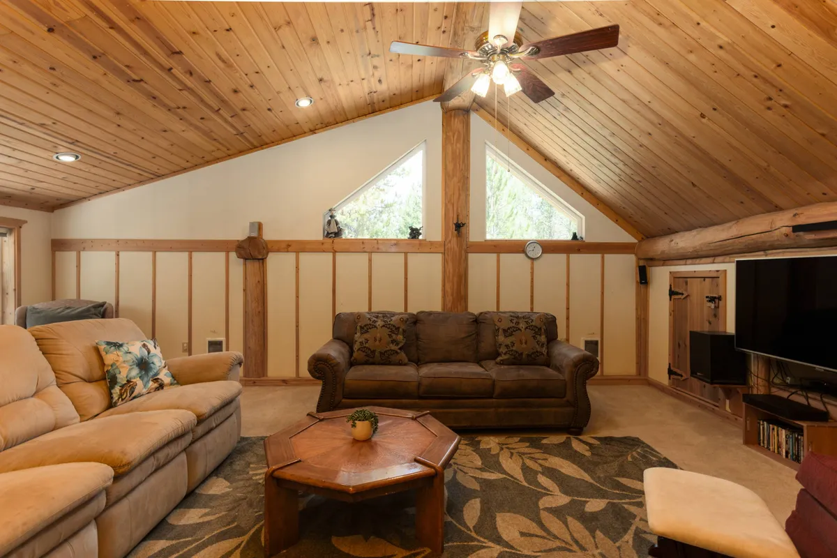 A cozy loft-like interior with exposed wooden beams, comfortable seating, a decorative clock, and a television setup, bathed in natural light. The arrangement invites both relaxation and entertainment, with space for small groups or individuals to enjoy a movie or lounge.