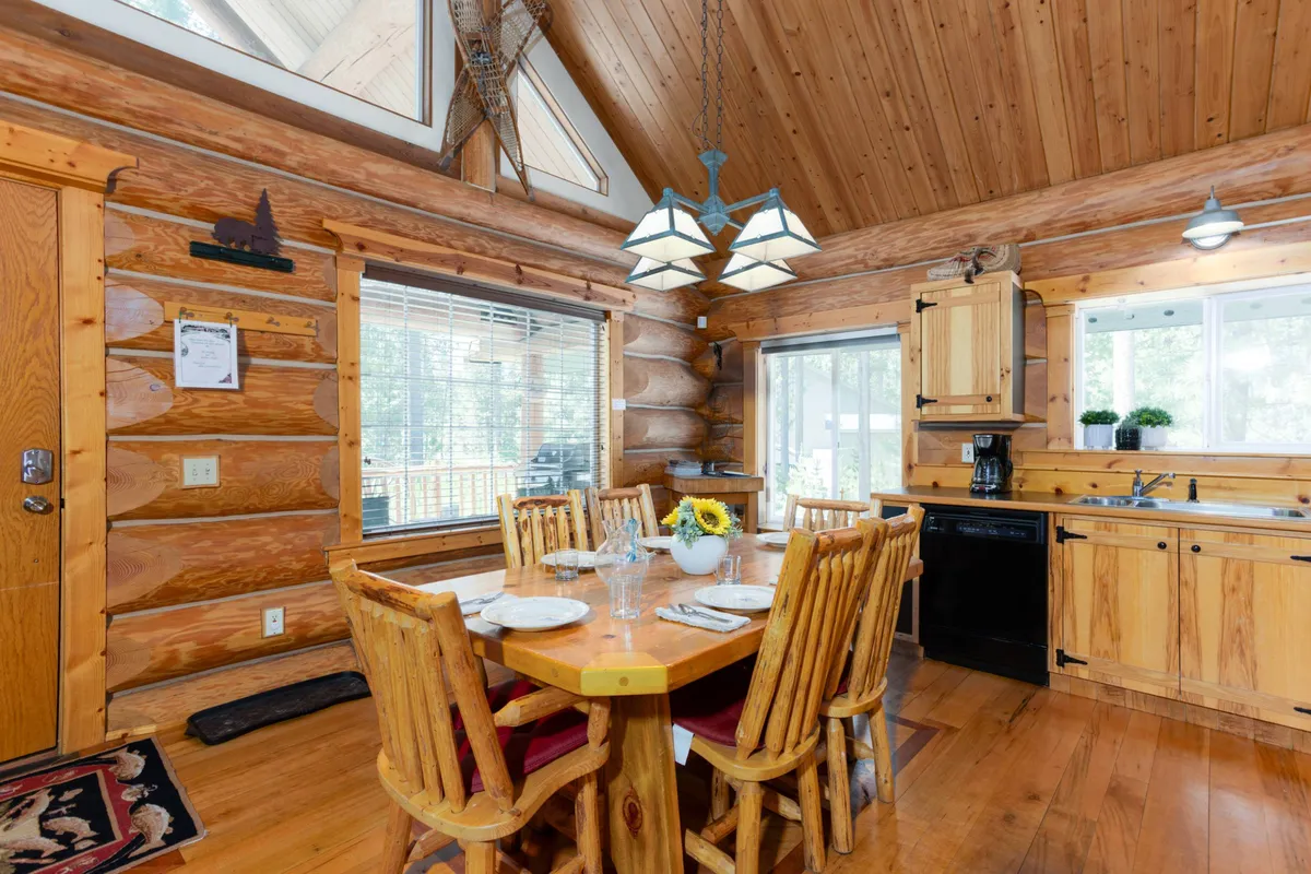 An inviting interior scene with a sturdy wooden dining table, log walls, large windows that fill the space with natural light, and a view of the adjacent kitchen area. The setting feels ready for family gatherings or intimate meals, with rustic and welcoming details throughout.