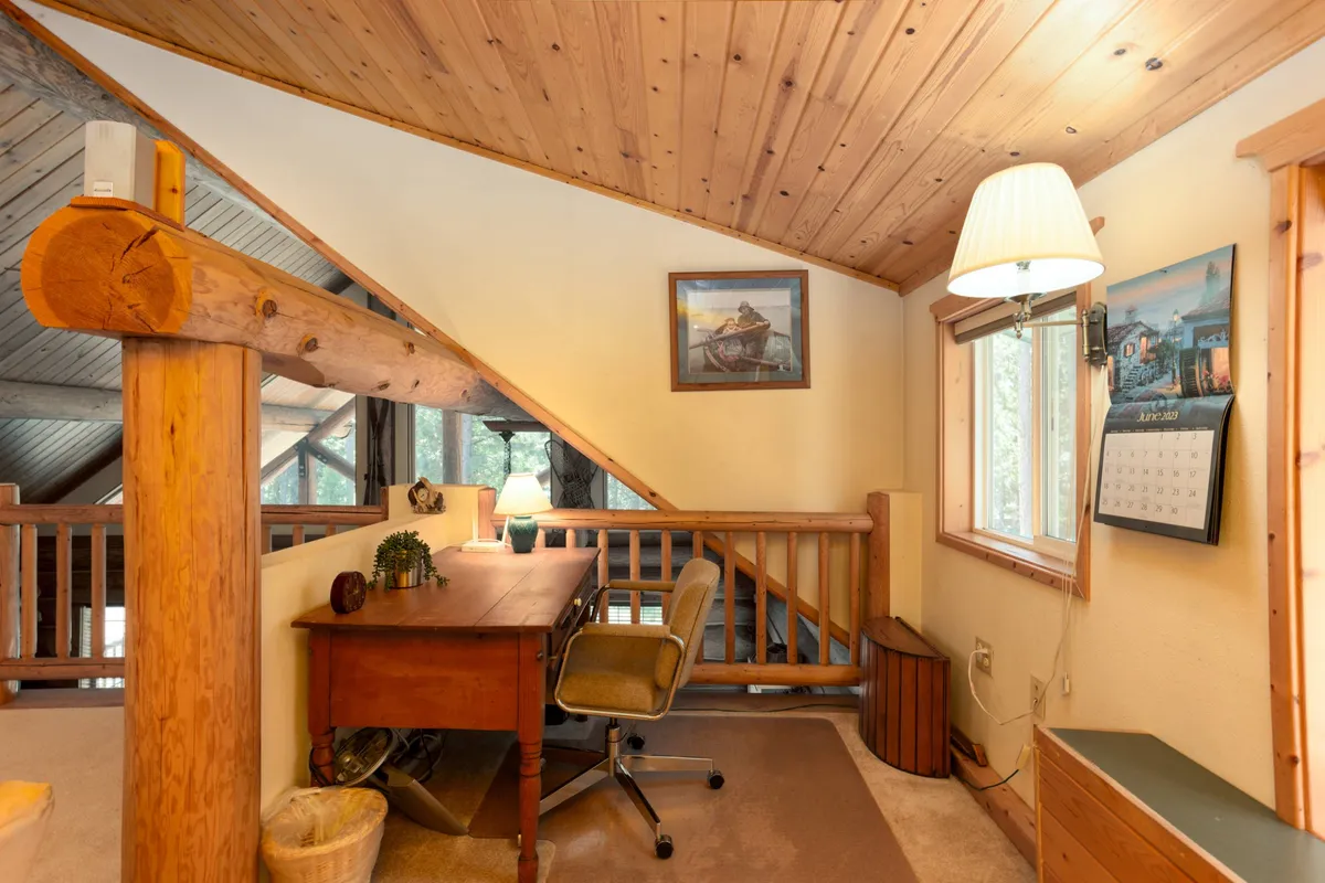 An upstairs loft area with a wooden desk positioned near a sunlit window, exposed beams, and tasteful wall art. The nook is set up for productive work or quiet reading, seamlessly blending work and leisure.