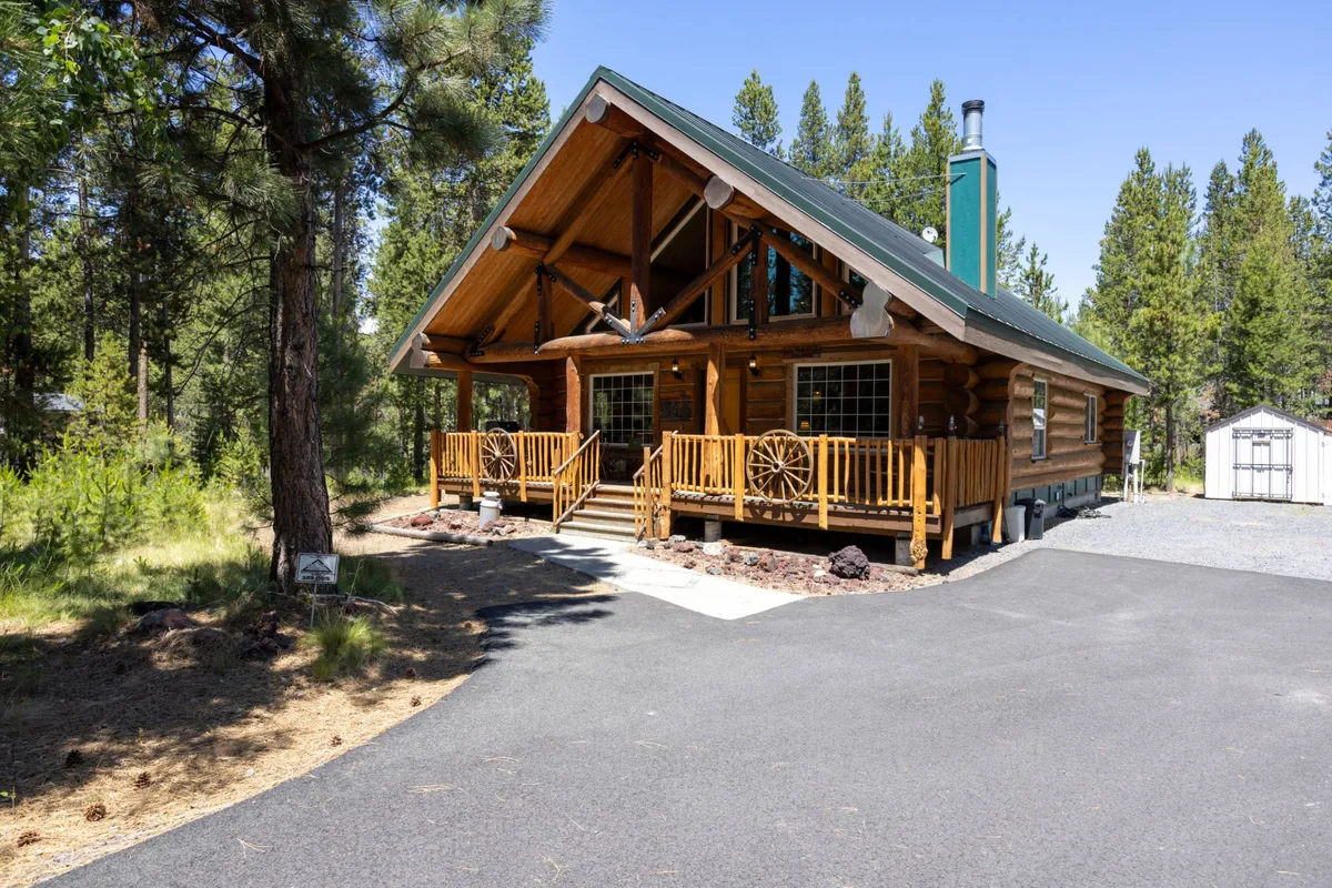 The exterior of a cozy log cabin with a classic front porch, wooden railings, and decorative wagon wheel. The setting is enveloped by tall trees, creating a sense of seclusion and connection to the outdoors—ideal for gatherings or a quiet evening under the stars.