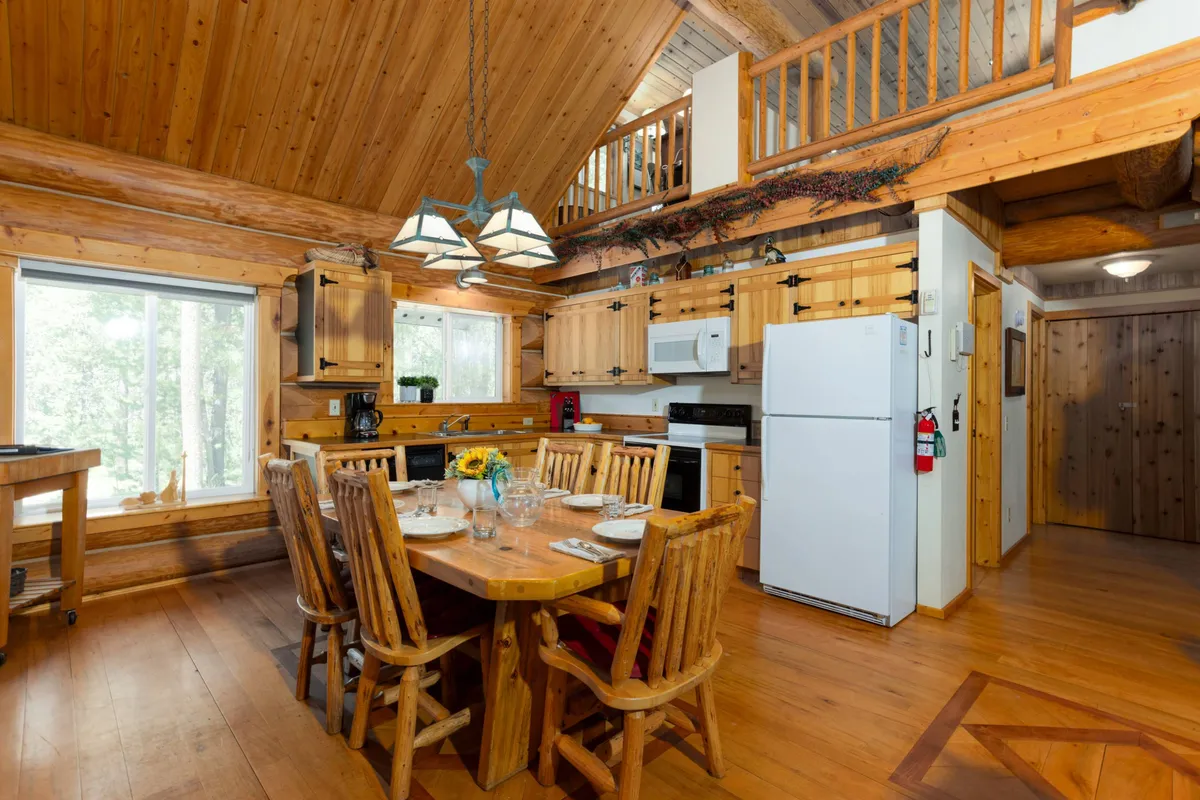 A sunlit open-plan interior featuring a wooden dining table at the center, a fully equipped kitchen with large windows, and exposed wooden beams that echo the cabin's rustic charm. The view through the windows hints at scenic outdoor vistas, while the setup encourages communal cooking and shared meals.