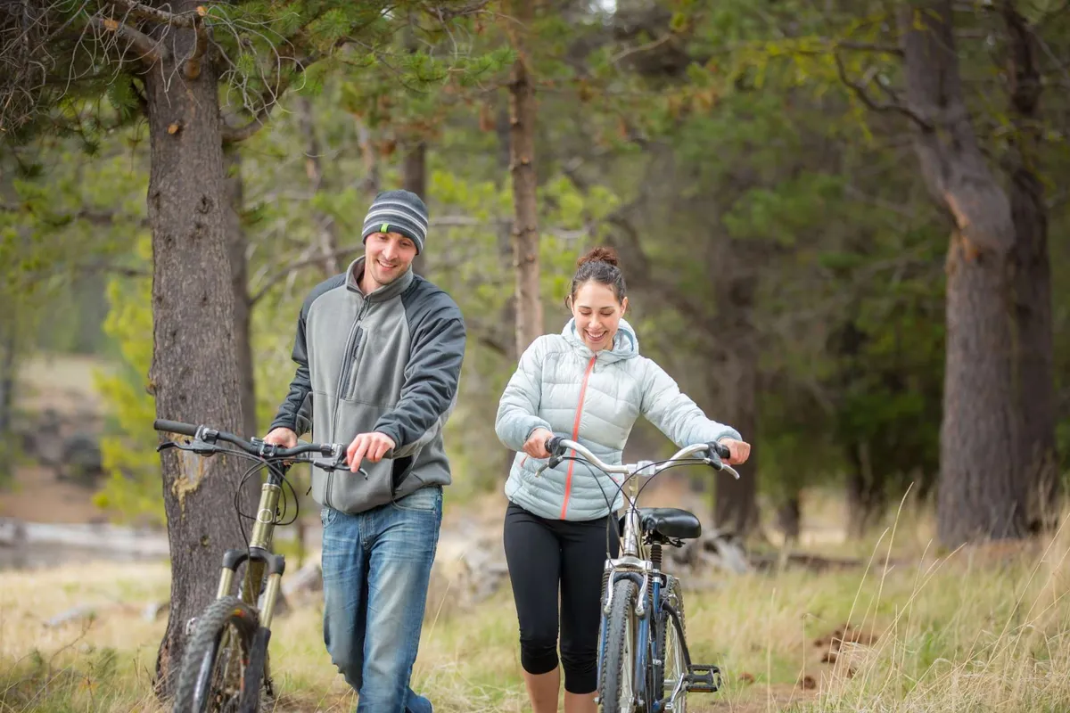An exterior image showing two cyclists walking with their bikes along a scenic path bordered by tall trees and open grassy areas. The scene exudes a spirit of camaraderie and enjoyment of the outdoors.