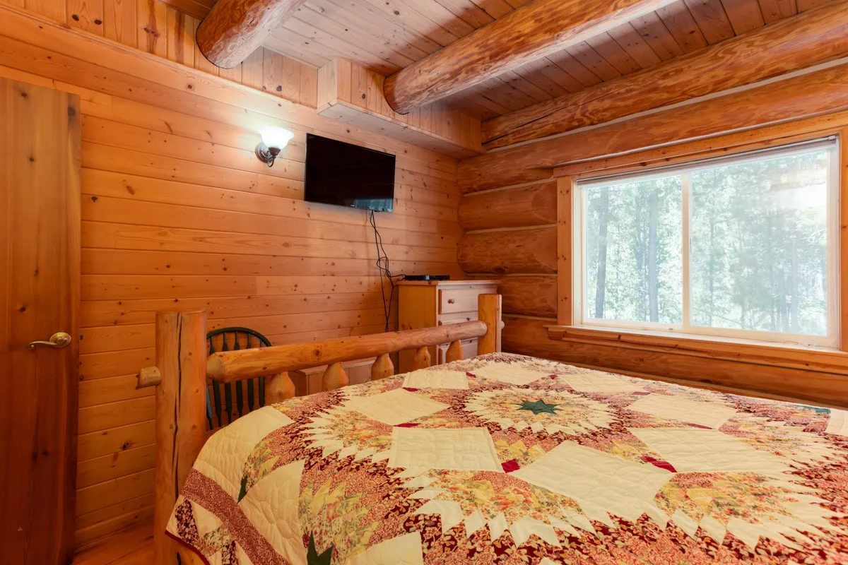 A warm, inviting bedroom featuring a wooden bed frame, a quilted bedspread, large window, and log walls. The space feels both private and homey, perfect for relaxation or enjoying peaceful moments with a view outside.