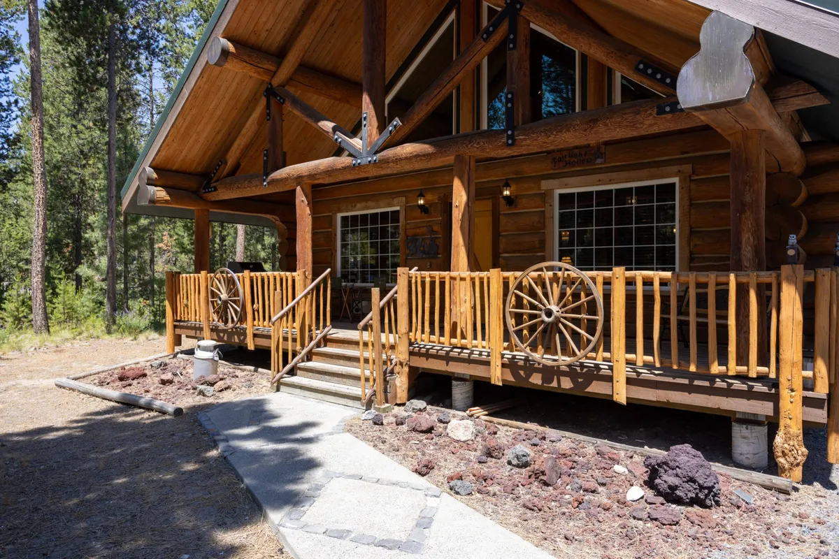 A charming log cabin structure with rustic wooden railings and wagon wheels sits amidst a natural landscape. A stone pathway leads to the porch, where guests can relax and enjoy the outdoors, with plenty of open space visible for activities.