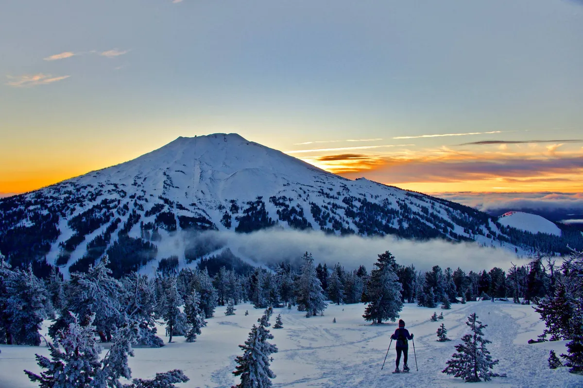 A breathtaking scene of a snow-covered mountain at sunset, with a hiker making their way along the ridge. The soft, golden light over the snowy landscape evokes both the thrill and tranquility of winter exploration.