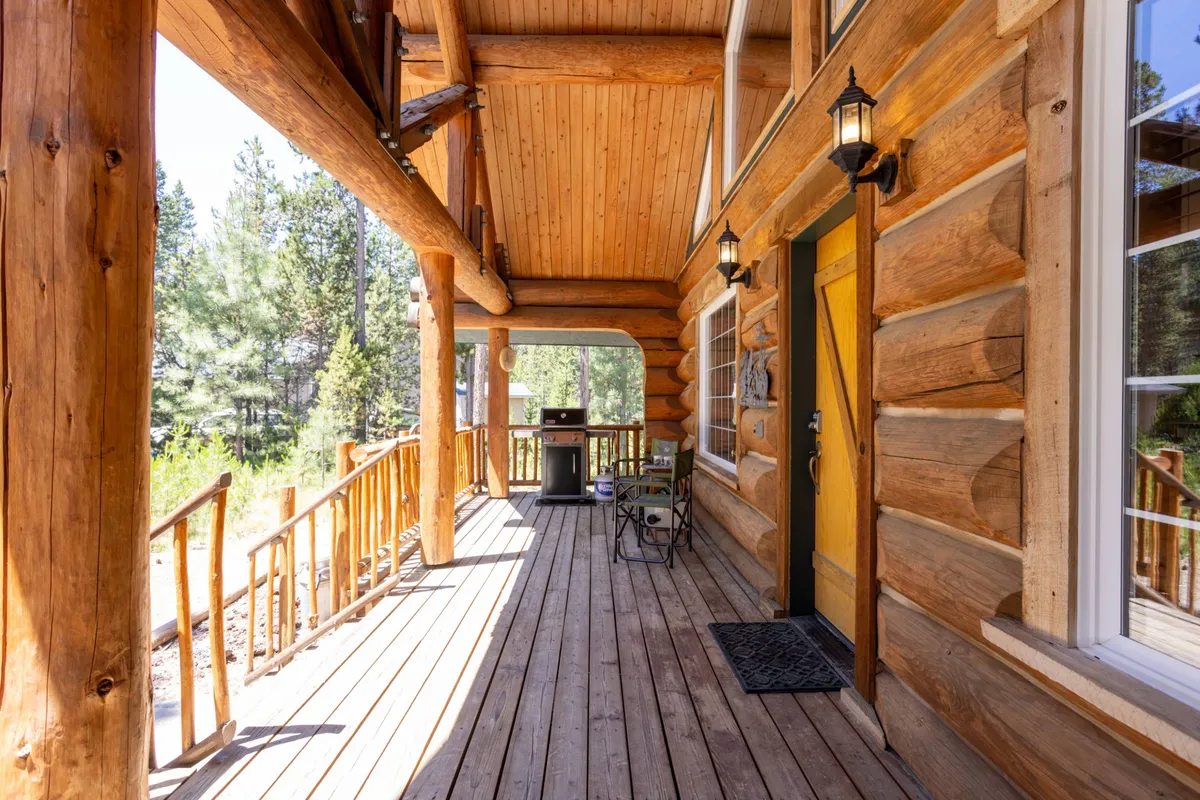 The exterior porch of the log cabin, featuring a distinctive yellow door, wooden railings, and an outdoor grill. The scene evokes cozy gatherings on the covered porch, with hints of sunset light and space for barbecuing or relaxing in nature.