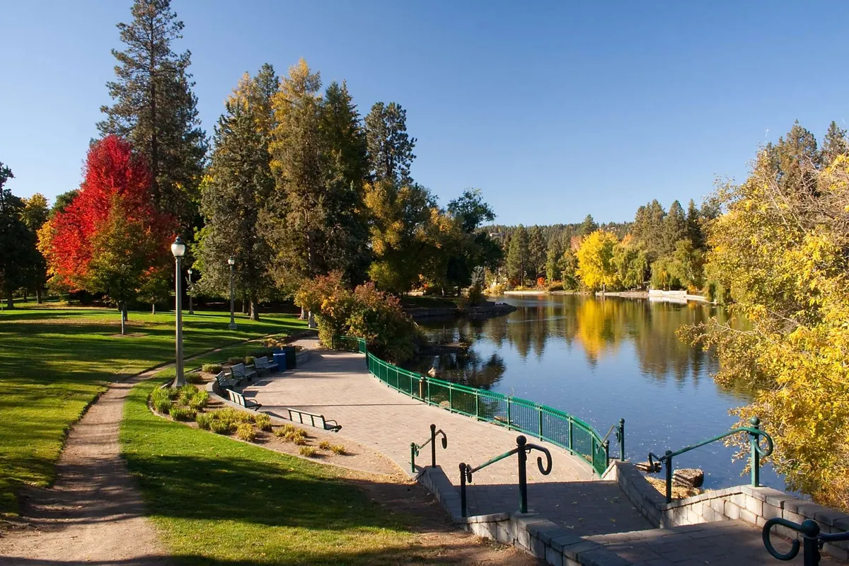 A scenic natural setting featuring calm waters bordered by colorful trees, with winding pathways and benches inviting relaxing walks, picnics, or birdwatching. The area evokes the accessible recreational amenities and tranquil environment found near Sunriver Resort.