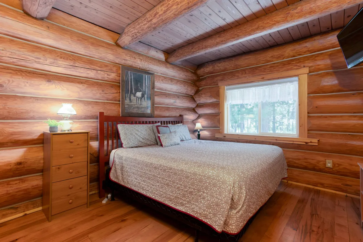A warmly lit bedroom with log cabin walls, a comfortable bed, a nightstand, and a window framing woodland views. The space feels inviting and tranquil, evoking the comfort and rustic charm described in the retreat narrative.
