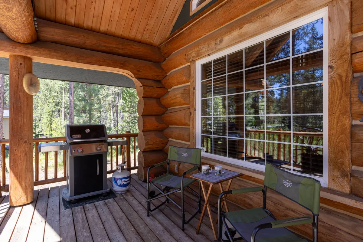 An inviting exterior shot of a covered outdoor space featuring a grill, table, and chairs, with a nearby window offering views into the cabin. The area is clearly designed for socializing, barbecuing, and relaxing—ideal for year-round use regardless of weather.